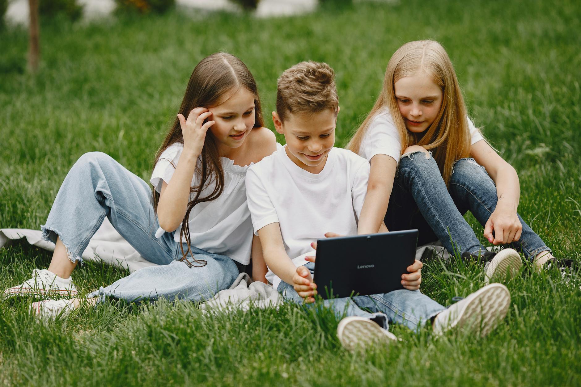 Three children using a tablet outdoors on a sunny day, sharing technology fun.